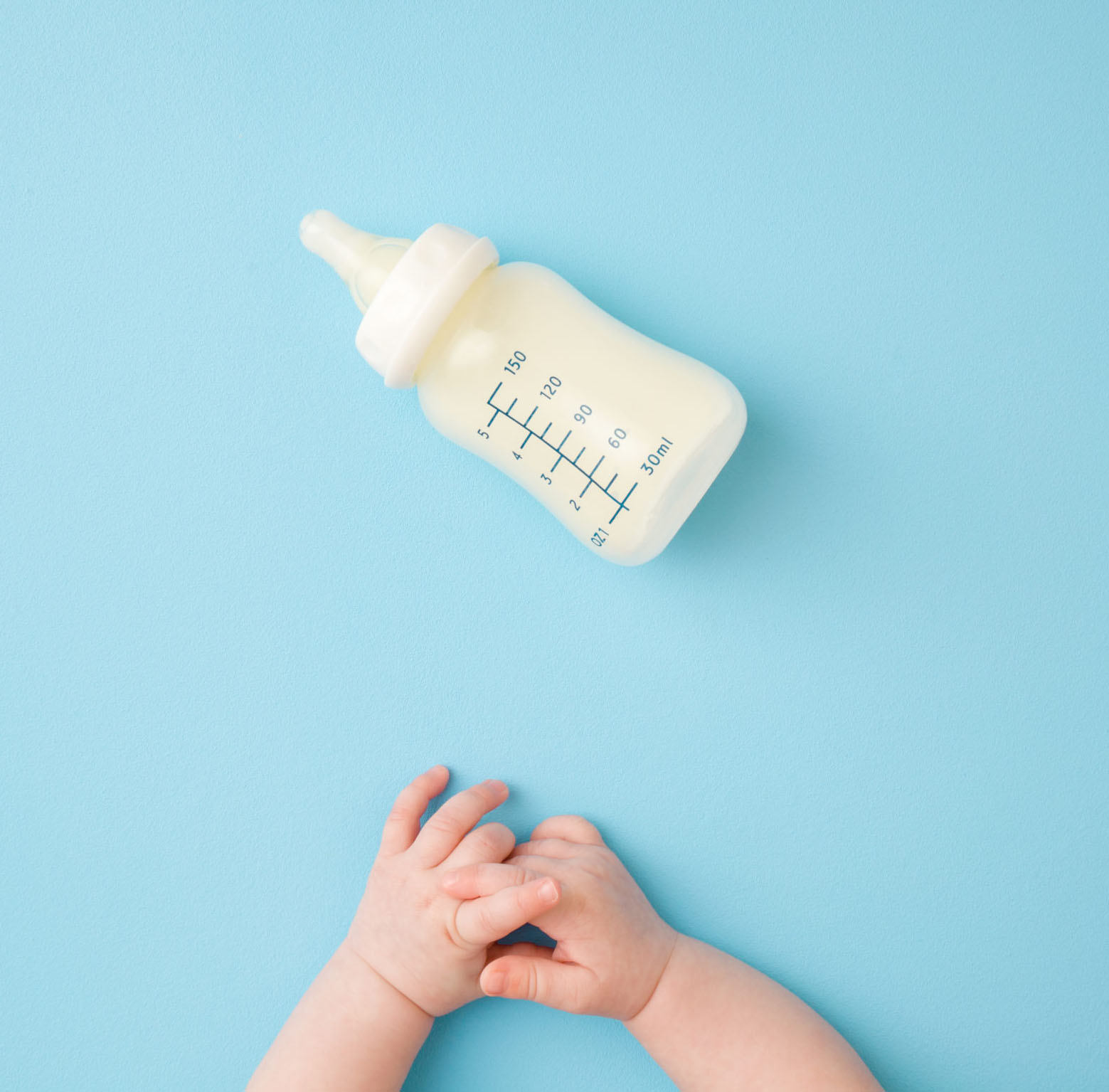 Infant hands and bottle of milk on light blue table background. baby's hands clasped on blue background with empty formula bottle